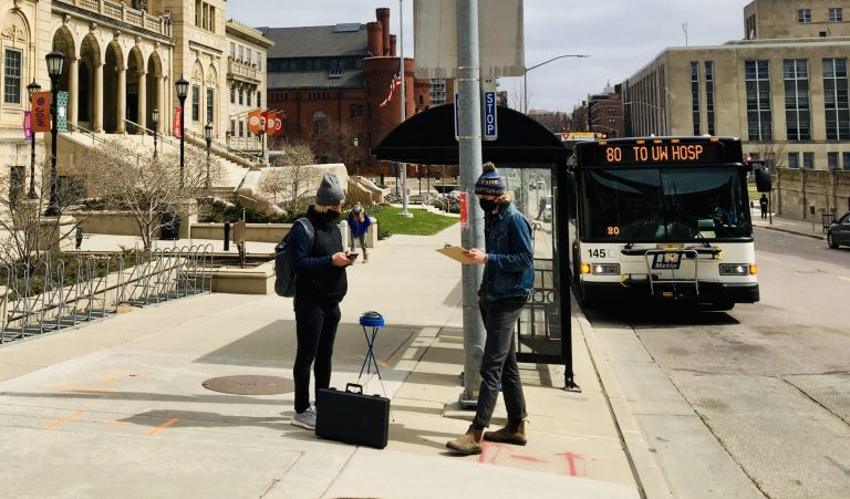 Two people standing on a sidewalk with a metro bus on the right and the Wisconsin Union building on the left