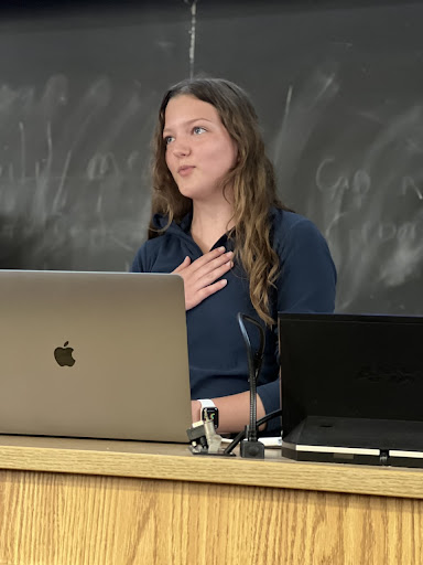 Caroline Crawley presents at a podium in a dark shirt, with a hand to her chest