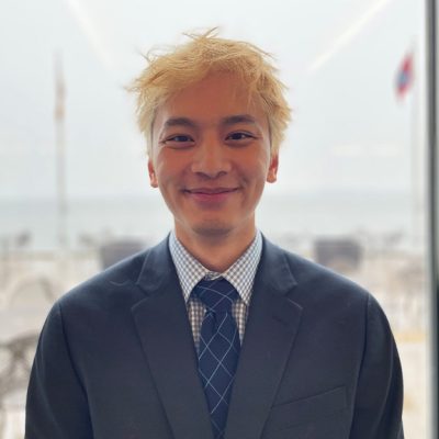 Young man with yellow-blonde hair and a dark blue suit in front of a light background