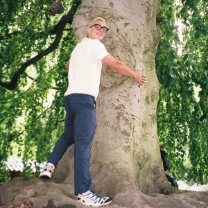 Young man in white tee shirt and blue pants hugging a tree while looking back at the camera and smiling