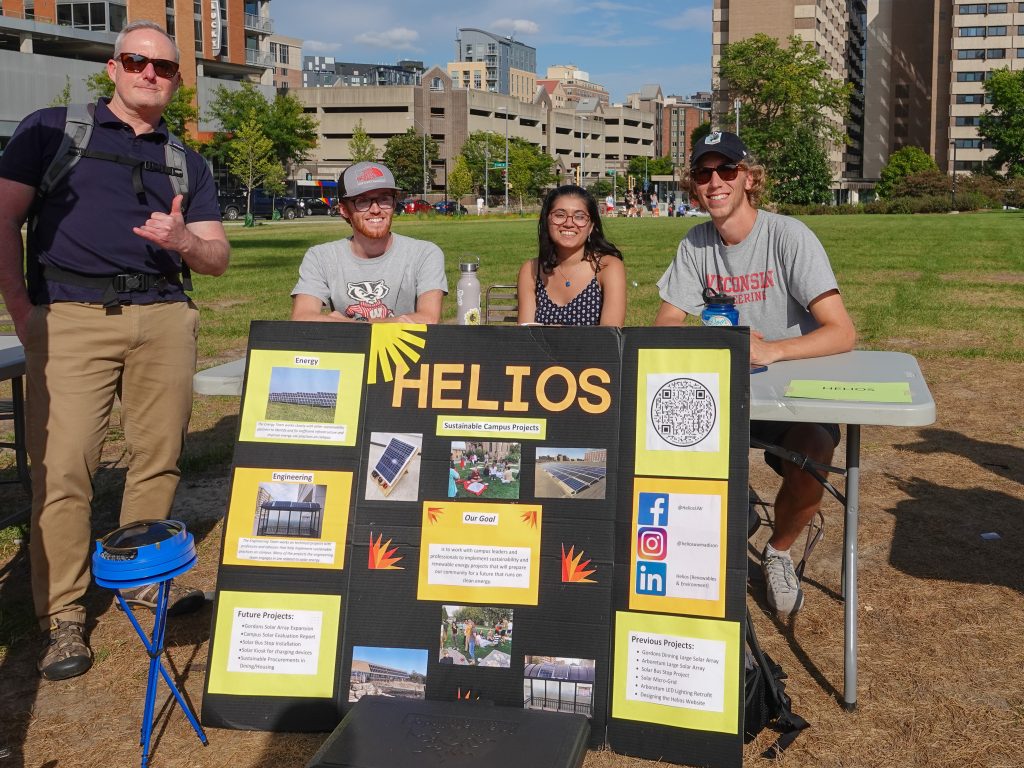 Black sign with "Helios" on it with four people behind it in front of a field and campus buildings