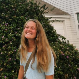 Young woman with long dark blond hair smiling widely in front of a flowering shrub