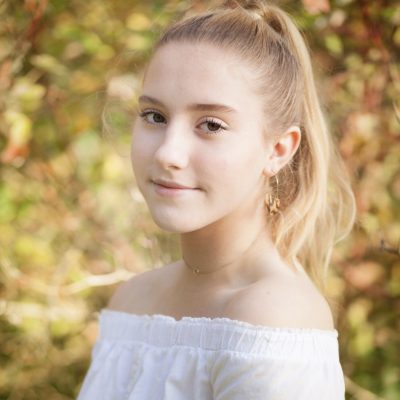 Young woman with blond hair in a ponytail looking over her shoulder at the camera with an autumnal background