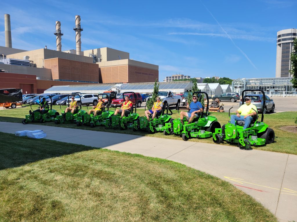 Several people sit on green lawn mowers on a sunny day