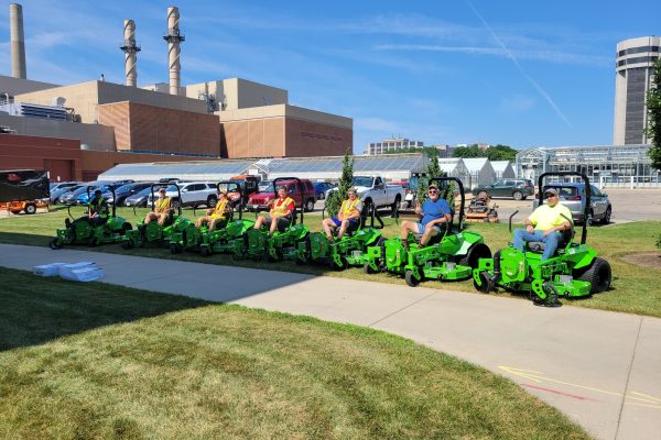 Several people sit on green lawn mowers on a sunny day