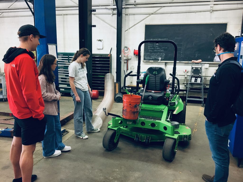 Four people standing around a green ride-on mower in a machine shop