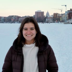 Carolyn Shumaker in a black coat and white shirt standing in front of a white frozen lake with the Madison skyline behind