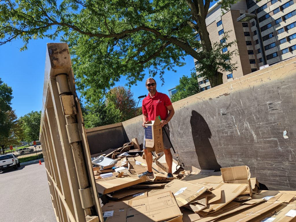 Travis Blomberg in a red shirt standing in an open dumpster on top of flattened cardboard boxes. 
