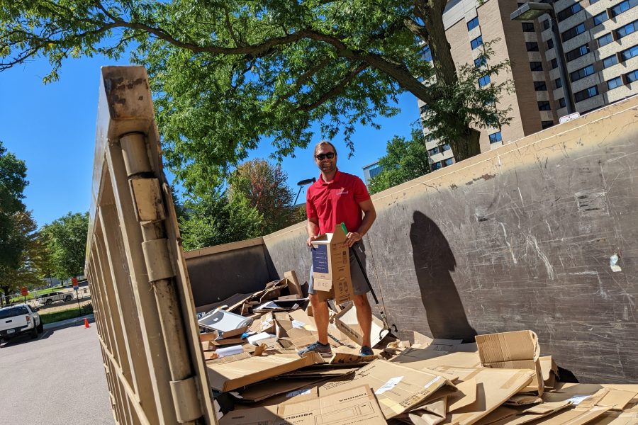 Travis Blomberg in a red shirt standing in an open dumpster on top of flattened cardboard boxes.